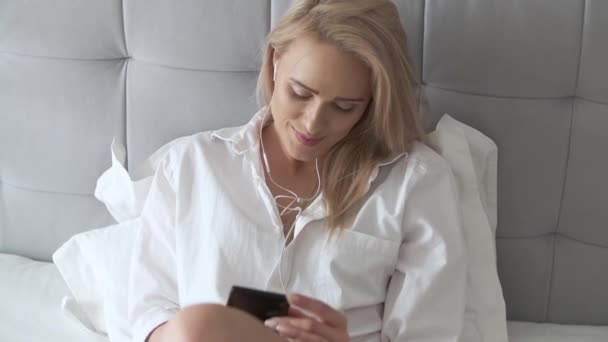 아름 다운, 웃는 금발 여자 흰 침대에 누워 및 smartphone을 사용 하 여 — 비디오
