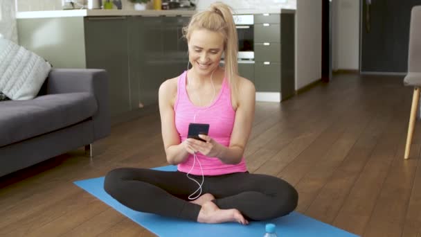 Carino donna seduta su tappetino yoga e la navigazione cellulare per le canzoni — Video Stock