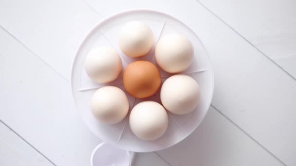 Ovos de galinha em um fogão elétrico de ovo em uma mesa de madeira branca — Vídeo de Stock