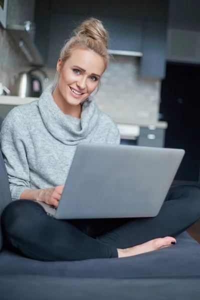 Cheerful young blond woman sitting on couch in living room and using laptop — Stock Photo, Image