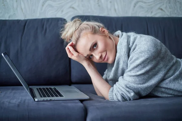 Mujer rubia feliz acostada propensa en el sofá y trabajando en el ordenador portátil —  Fotos de Stock