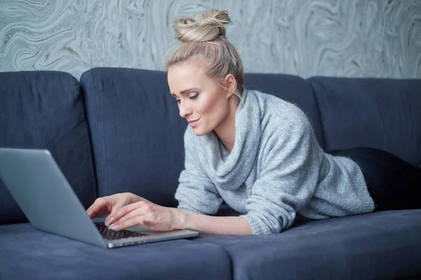 Gelukkige blonde vrouw liggend vatbaar op sofa en werken op laptop computer — Stockfoto