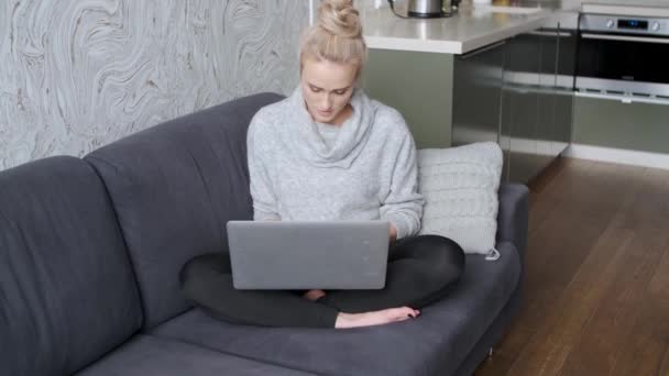 リビングルームでソファに座って、ラップトップを使用して陽気な若いブロンドの女性 — ストック動画