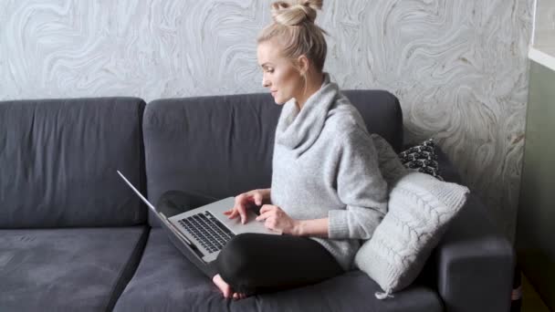 Vrolijke jonge blonde vrouw zittend op de Bank in de woonkamer en het gebruik van laptop — Stockvideo