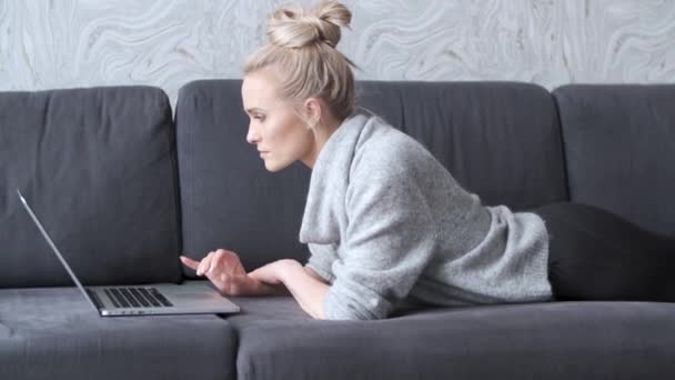 Gelukkige blonde vrouw liggend vatbaar op sofa en werken op laptop computer — Stockvideo