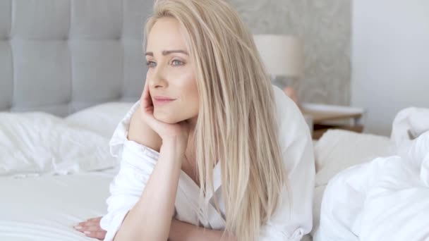 Hermosa mujer de mediana edad acostada en la cama, vistiendo camisa blanca y sonriendo — Vídeos de Stock