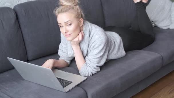 Glückliche blonde Frau liegt geneigt auf Sofa und arbeitet am Laptop — Stockvideo