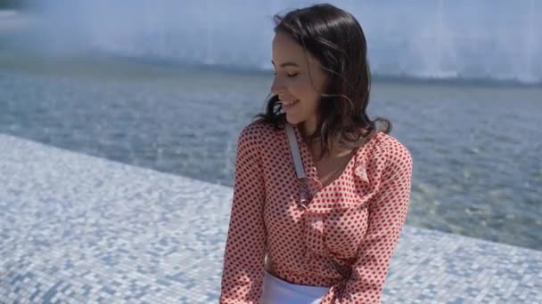 Portrait de belle fille en vêtements d'été colorés assis près d'une fontaine — Video