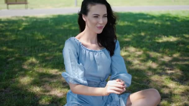Porträt einer attraktiven brünetten Frau im blauen Kleid, die in einem Park sitzt — Stockvideo