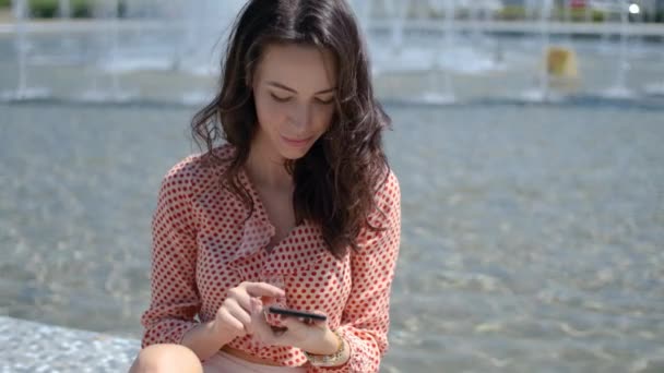 Adorable, mignonne femme brune, assise à côté de la fontaine de la ville en été — Video