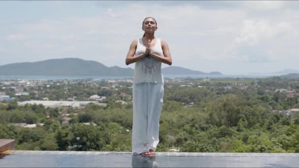 Mulher meditando em pé junto à piscina contra a costa tropical — Vídeo de Stock