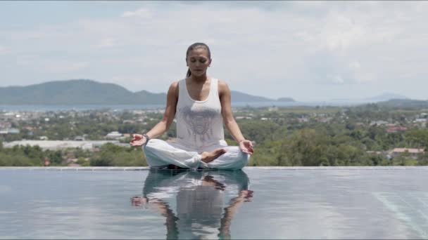 Mujer tranquila practicando yoga en postura de loto junto a la piscina contra la ciudad turística — Vídeos de Stock