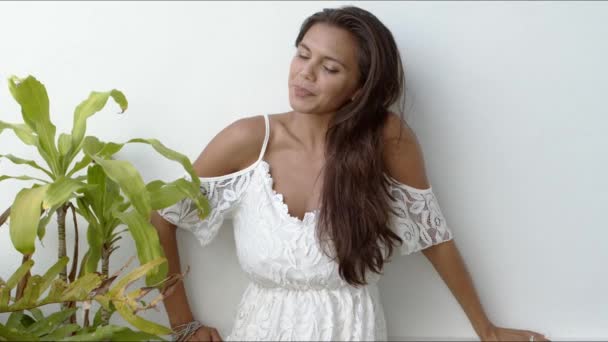 Счастливая брюнетка в белом платье стоит рядом с зеленым растением — стоковое видео