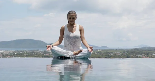 Femme sereine pratiquant le yoga en position lotus au bord de la piscine contre le ciel — Photo