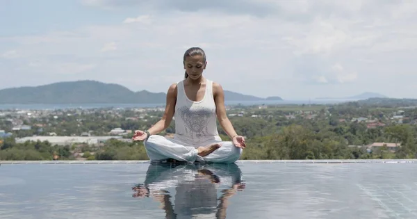 Calm woman practicing yoga in lotus posture poolside against resort town Stock Photo