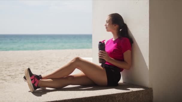 Sportos nő üveg vízzel pihentető edzés után a strandon — Stock videók