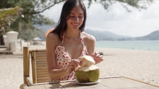 Młoda kobieta siedzi przy stole z kokosem na plaży — Wideo stockowe