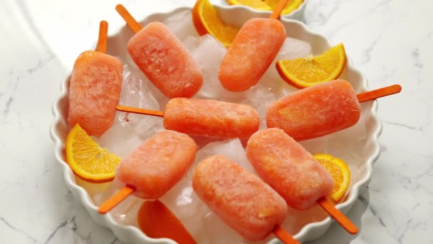 Homemade, juicy, orange popsicles. Placed on a white plate with ice cubes — Stock Video