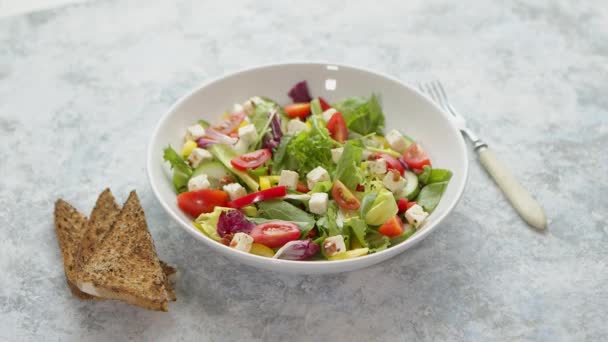 Красочный свежий овощной салат с сыром фета в белой глубокой миске крупным планом на каменном столе — стоковое видео