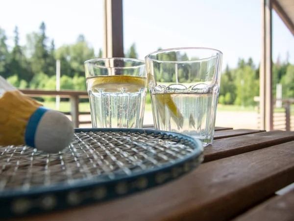 Wooden country furniture on the terrace of a country house. Badminton rackets and shuttlecock on wooden table. Two glasses with refreshing lemonade in the background. Lush green foliage in the
