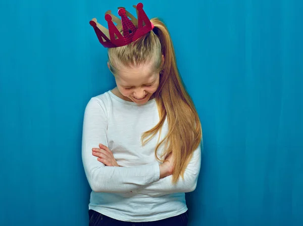 Little girl wearing pink crown laughing while standing with crossed arms on blue wall background