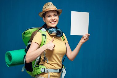 Sırt çantasıyla gezmeye hazır gülümseyen bir kadın küçük bir tabela tutuyor ve baş parmağını gösteriyor..