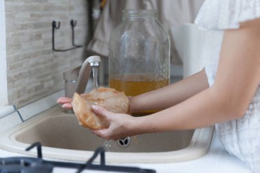Young woman preparing her kombucha tea in the kitchen.She is cleaning the kombucha in cold water. clipart