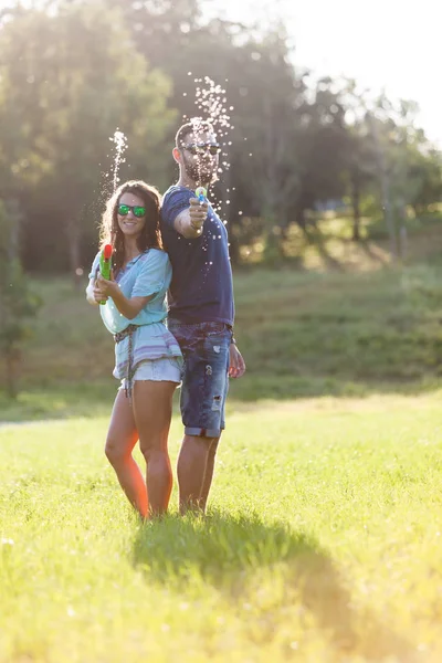 A playful young couple chasing each other and playing with water guns in a meadow ar sunset.