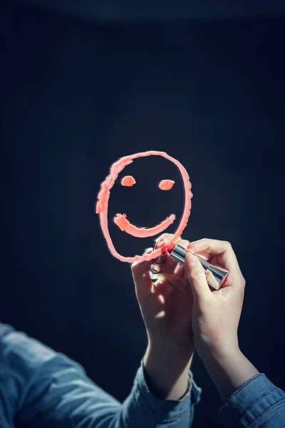 Woman drawing smiley face on mirror with red lipstick.