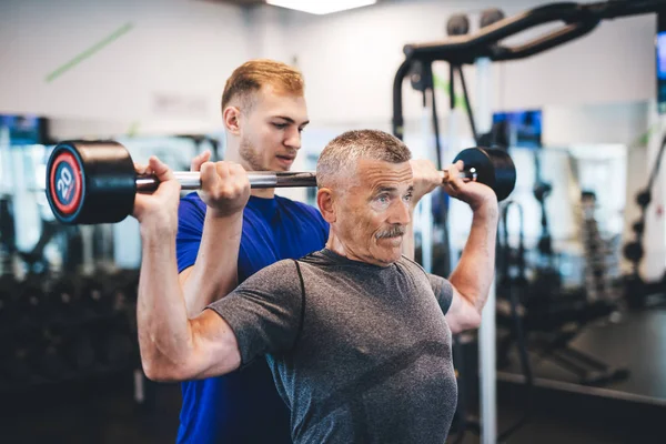 Senior man lifting weights with help of gym assistant. Work of personal trainer. Bodybuilding.