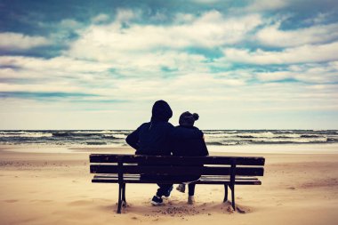 Back view of father and daughter sitting on bench on sandy beach clipart