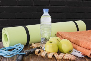 Yoga Mat ve su şişesi yakın çekim. Spor malzemeleri, üç yeşil elma ve bir ip atlama. Siyah tuğla duvara karşı.