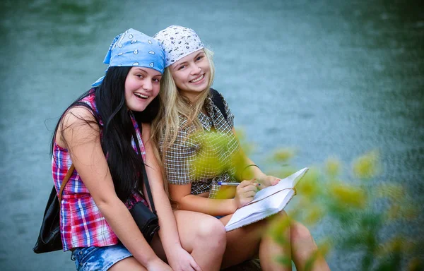 二人の幸せな女性友人ノートが付いている湖の近くの石の上に座ってバックパック — ストック写真