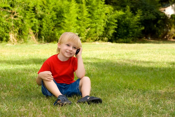 快乐的小男孩坐在公园的绿草上谈论手机 — 图库照片
