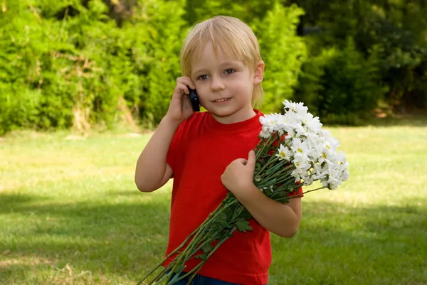 快乐的小男孩捧着白花 电话站在绿草的背景上说话 — 图库照片