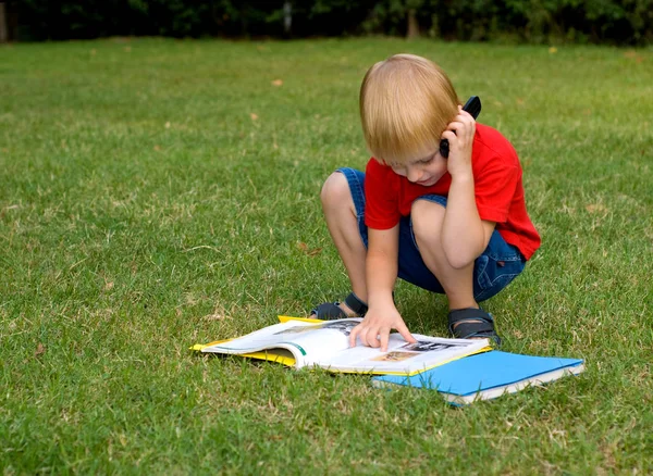 かわいい男の子本で緑の草の上に座って携帯電話で話して — ストック写真