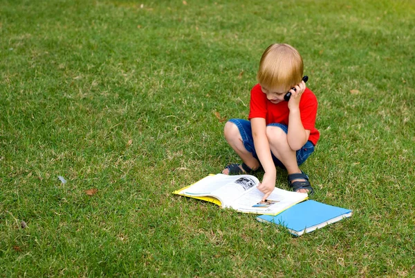 かわいい男の子本で緑の草の上に座って携帯電話で話して — ストック写真