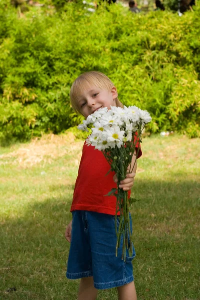 緑の草の背景に立っている白い花を持って幸せの小さな男の子 — ストック写真