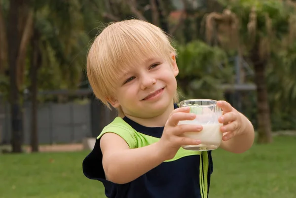 户外肖像的快乐小男孩拿着玻璃牛奶站在模糊的绿色背景 — 图库照片