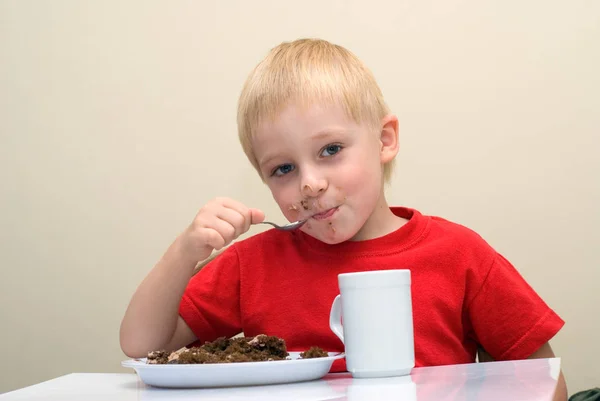 快乐的小男孩吃自制的蛋糕坐在桌上用勺子和茶杯 — 图库照片