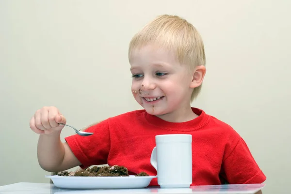 快乐的小男孩吃自制的蛋糕坐在桌上用勺子和茶杯 — 图库照片