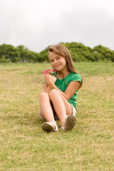 緑の草の完全な長さの肖像画の上に座ってのピンクの花を持って幸せな少女 — ストック写真