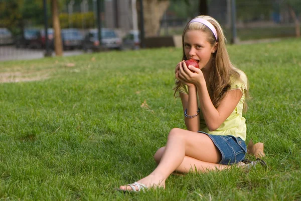 公園内の緑の草の上に座って赤いリンゴを保持しているかわいい十代の少女の屋外のポートレート — ストック写真