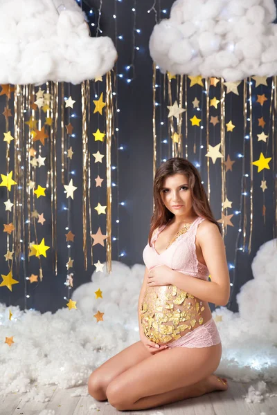 Mujer embarazada Glamour en un estudio de Navidad con una guirnalda de estrellas de oro, nubes esponjosas y luces — Foto de Stock