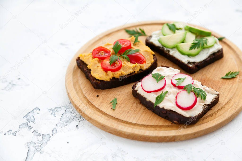 Close-up tasty vegetarian rye bread toasts with cottage cheese, hummus, avocado, raddish and tomato. Wooden board on a white concrete background