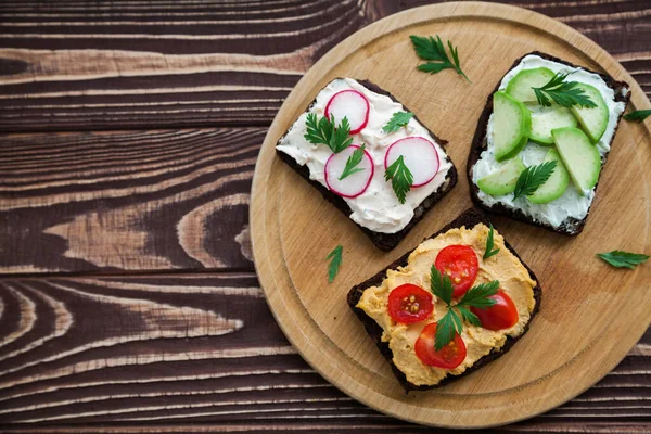 Vegetarian rye bread toasts with cottage cheese, hummus, avocado, raddish and tomato. Wooden board on a rural background, top view, copy-space