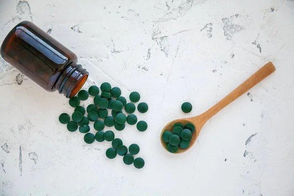フラット 緑のらせん状の丸薬のボトルの白い具体的な背景にドロップされます 木製のスプーンでいくつかの錠剤 スーパーフードのコンセプト スピルリナの栄養補助食品 — ストック写真