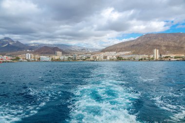 Tenerife 'nin canlı kıyı kasabası Los Cristianos' un denizinden bir görüntü, önplanda güçlü bir tekne, dağların manzarası ve hareketli bir liman