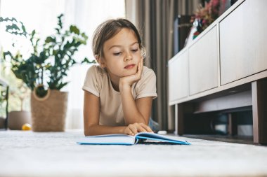 Küçük bir kız yerde uzanıyor, kitap okurken derinden odaklanmış. Bu fotoğraf öğrenme, hayal gücü ve rahat bir evde, eğitim ve çocukluk gelişimi temaları için mükemmel boş zamanları temsil ediyor.