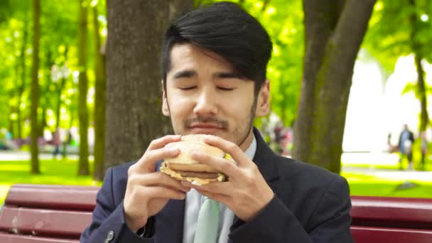 アジア系のビジネスマンは公園とハンバーガーをスニッフィングのベンチに座っています。 — ストック動画